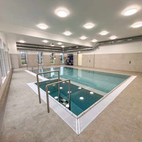 Schwimmbecken mit zwei Zugängen mit Metallgeländer. Runde Deckenleuchten spiegeln sich im Wasser. Im Hintergrund sind eine Einstieghilfe (ein schwenkbarer Sitz) für gehbeeinträchtige Personen sowie Schwimmutensilien zu sehen.